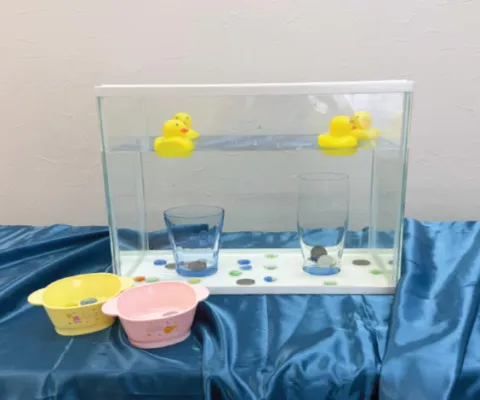 水を張った水槽にアヒルが浮かび、底にコインとコップがあり、手前におわんが置いてある