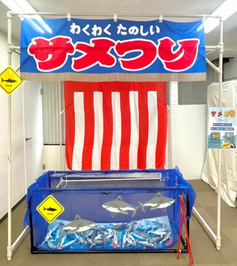 サメ釣りの屋台。紅白幕の前に、サメのフィギュアが入った水槽がある